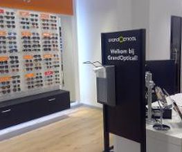 Winkelinterieur met een zonnebrillenwand en balie bij GrandOptical, met een Welkomstbord.