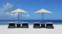 Strand mit vier Liegen und zwei Sonnenschirmen am Meer unter blauem Himmel.
