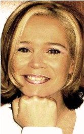 Blonde Frau mit kurzem Haar lächelt in die Kamera, stützt Kinn auf Hand. Hintergrund unscharf.