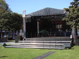 Leere Bühne im Freien mit schwarzem Dach, umgeben von Bäumen an einem sonnigen Tag.