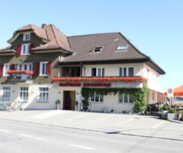 Zweistöckiges Haus mit rotem Schild und Terrasse, Fahrräder davor geparkt, sonniger Tag.
