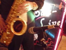 Mann spielt Saxophon auf einer Bühne, Mikrophon im Vordergrund, unscharfes Live-Schild im Hintergrund.