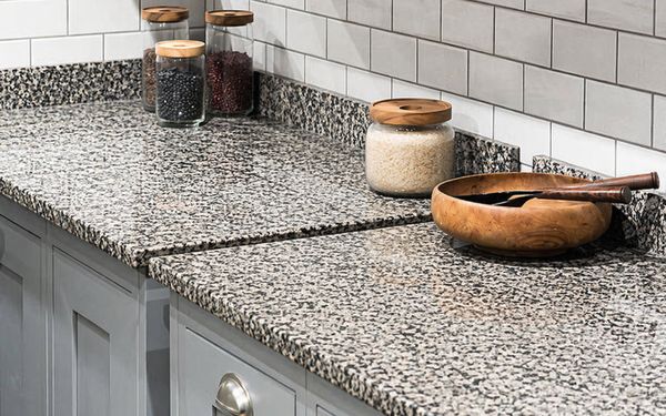 Encimera de cocina de granito con patrón moteado en tonos gris y negro, superficie pulida, acompañada de tarros de vidrio con alimentos y un bol de madera sobre muebles blancos y salpicadero de azulejos.