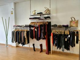 Ropa colgada en una tienda, estantes con bolsos y pantalones, suelo de madera.