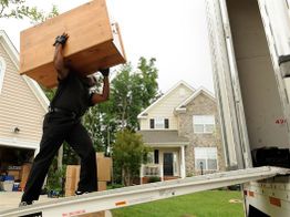 Person lifting a large box into a moving truck near a suburban house.