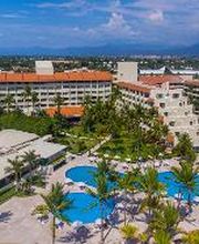 Vista aérea de un resort con piscinas azules y palmeras, rodeado de edificios y montañas al fondo.