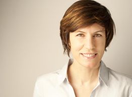 Eine Frau mit kurzem braunem Haar lächelt in die Kamera, trägt ein weißes Hemd.