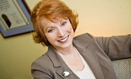 Smiling person with red hair in a suit, sitting in an office with a diploma on the wall.