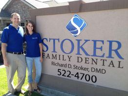 Two people in blue shirts by a "Stoker Family Dental" sign on a sunny day.