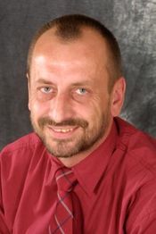 Eine lächelnde Person mit Bart, in rotem Hemd und Krawatte vor grauem Hintergrund.