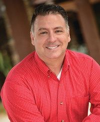 Smiling man in a red checkered shirt, sitting outdoors with a blurred background.
