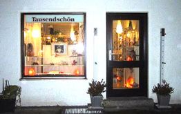 Beleuchtetes Schaufenster und Eingang eines Geschäfts namens "Tausendschön" mit Dekorationen.