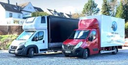Zwei Lieferwagen, ein weißer und ein roter, parken auf einem Kiesplatz vor Häusern und Bäumen.