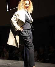 Frau auf Laufsteg in hellem Mantel und dunklem Anzug unter Bühnenlicht.