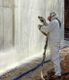 Worker in protective gear spraying a substance onto a large concrete wall.