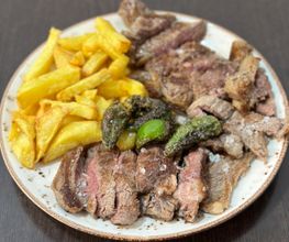Plato de carne asada con papas fritas y pimientos verdes sobre una mesa oscura.