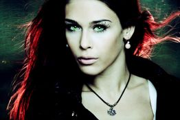 Frau mit roten Haaren und grünen Augen, trägt Ohrringe und eine Halskette, schaut ernst.