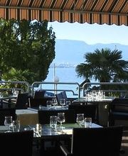 Restaurantterrasse am See mit gedeckten Tischen, Sonnenschirm, Bäumen und Bergblick im Hintergrund.