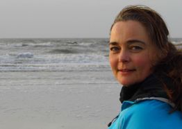 Eine Frau steht am Meer und blickt in die Kamera, mit windigem Haar und blauer Jacke.