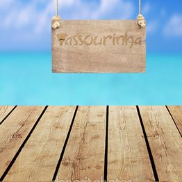 Placa de madeira pendurada sobre piso de tábuas, com mar azul e céu ao fundo.