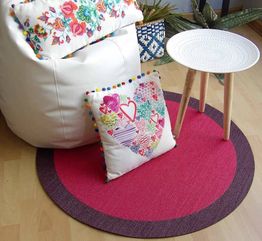 Sillón blanco con cojines coloridos, alfombra roja circular y mesa blanca sobre piso de madera.