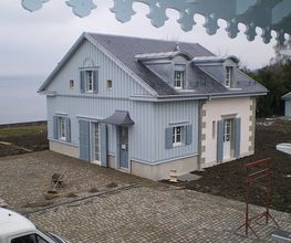 Ein graues Haus am Wasser, umgeben von Pflastersteinen und einer Rasenfläche, grauer Himmel.