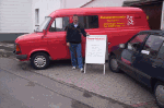 Ein Mann steht neben einem roten Lieferwagen mit einem Werbeschild auf dem Gehweg.