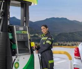 Mujer en gasolinera con uniforme, llenando combustible en coche blanco frente a montañas.