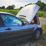 Car with open hood on roadside, person checking engine, green fields in background.