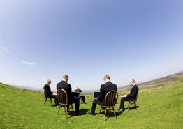 Vijf mannen in pakken zitten buiten op stoelen in een cirkel op een grasveld tegen een heldere hemel.