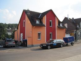 Zweifarbiges Haus in Orange und Rot mit Autos und Anhänger davor, auf einer ruhigen Straße.