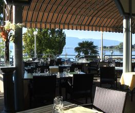 Überdachte Terrasse mit Tischen und Stühlen, Blick auf See und Berge im Hintergrund.