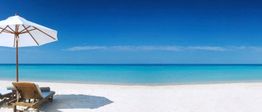 Strand mit weißem Sand, Sonnenschirm und Holzliegen, blaues Meer und klarer Himmel im Hintergrund.