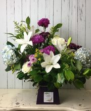 Ramo de flores con lirios blancos y flores moradas sobre una mesa. Fondo de madera clara.