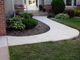 Curved concrete walkway with plants and shrubs leading to a house entrance.