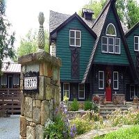 Green and black house with red door, stone pillar, and flowers in front yard.