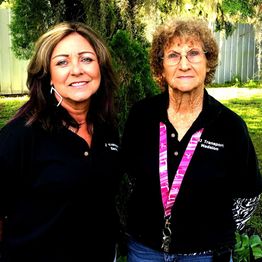 Two women in matching black shirts, standing outside near trees and a fence, smiling slightly.