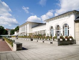Helles, modernes Gebäude mit großer Terrasse und Blumen, unter blauem Himmel mit wenigen Wolken.
