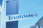 Schild mit der Aufschrift "Ersatzteile" vor blauem Himmel, rechts ein Gebäude.
