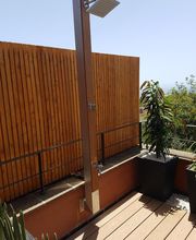 Ducha exterior en terraza con madera, plantas en macetas y cielo despejado.