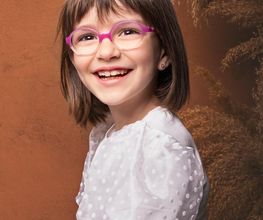 Niña sonriente con gafas rosas y blusa blanca, fondo marrón.