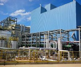 Usina industrial com estruturas metálicas, tubulações e prédio azul ao fundo, céu azul claro.