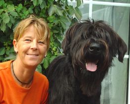 Frau in orangefarbenem Shirt lächelt neben einem fröhlichen, großen schwarzen Hund vor grünen Blättern.