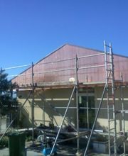 Gerüst an einem Hausdach unter blauem Himmel, Baum links, einige Werkzeuge auf der Baustelle.