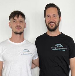Zwei Männer in weißen und schwarzen T-Shirts mit "Umzugsmeister GmbH"-Logo stehen nebeneinander.