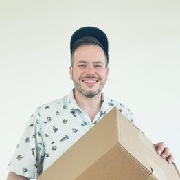 Ein Mann mit Cap hält einen großen Karton und lächelt in die Kamera.