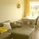 Wohnzimmer mit grauem Sofa, gelben Kissen, Esstisch, Stühlen und großen Fenstern im Hintergrund.