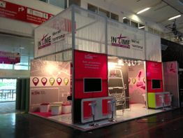 Messestand mit Monitoren, Hockern und Werbeplakaten in rosa und weiß, in Ausstellungsraum.