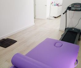 Sala blanca con camilla morada y caminadora; puerta abierta al fondo con vista al pasillo.