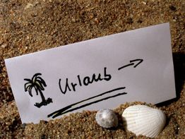 Notiz im Sand mit Palmenzeichnung und "Urlaub"-Schrift. Muscheln daneben.